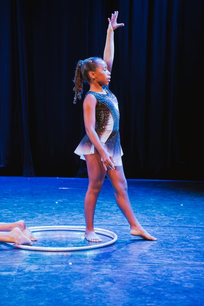 Young girl in a sparkly blue dance costume stands on stage with her right arm raised, a hoop at her feet.