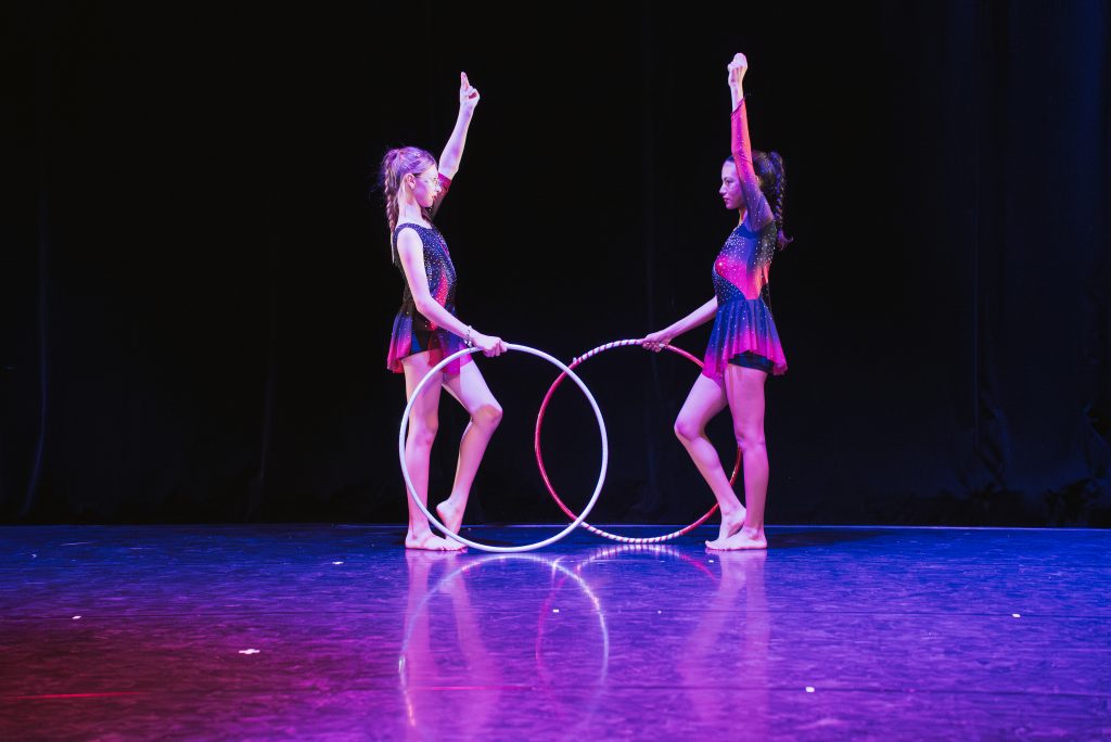 Two young female performers in matching sparkly costumes hold hoops on a dark stage, facing each other with one arm raised in a synchronized pose.