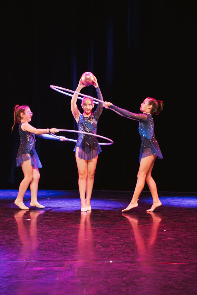Three young girls in sparkly blue costumes perform rhythmic gymnastics on stage with hoops and a pink ball.