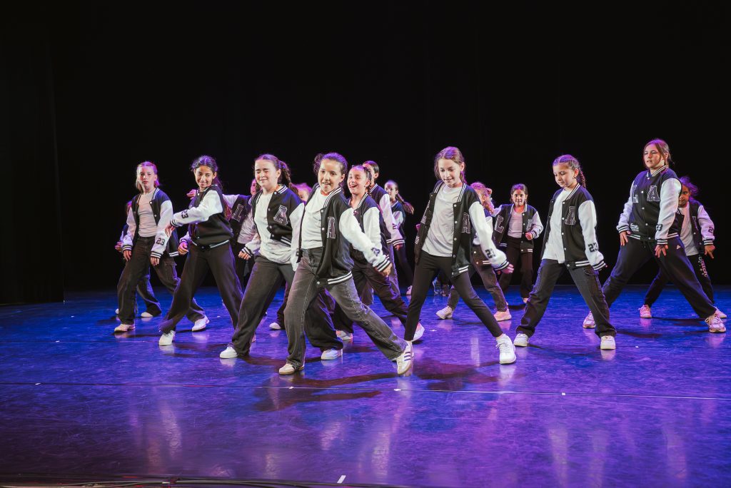Group of young dancers in matching black and white varsity jackets performing a synchronized routine on a dark stage with blue lighting.
