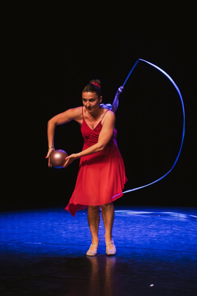 Female dancer in a red dress on a dark stage, spinning a blue hoop while holding a metallic ball