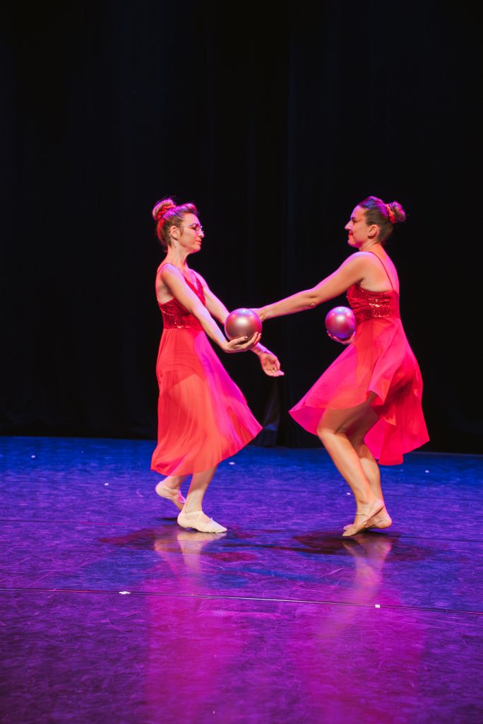 Two female dancers in bright pink costumes hold pink balls and face each other on a dark stage, mid-performance with purple lighting.