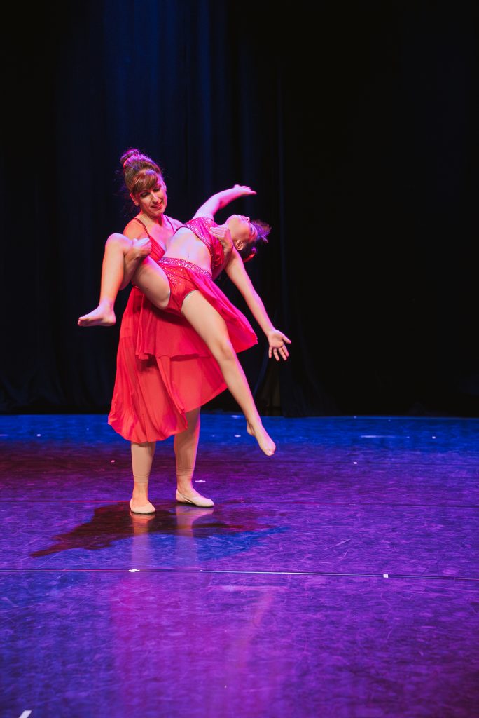 Two female dancers perform a lift on stage, one holding the other in a pink costume against a dark background.