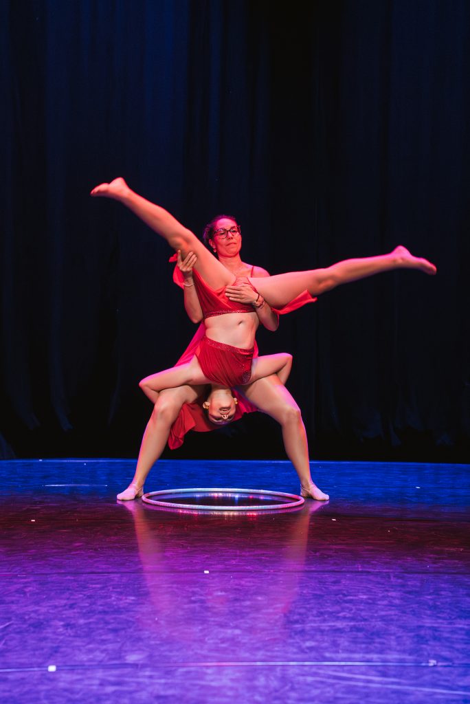 Dancers in red costumes perform an overhead split with a hoop on stage, black curtains behind them, purple lighting.