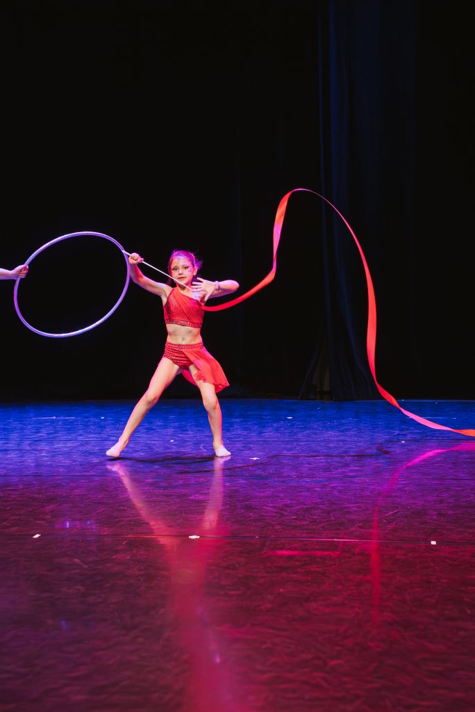 Young girl in a red costume performing on a dark stage with a purple hoop on her left and a flowing red ribbon on the right.