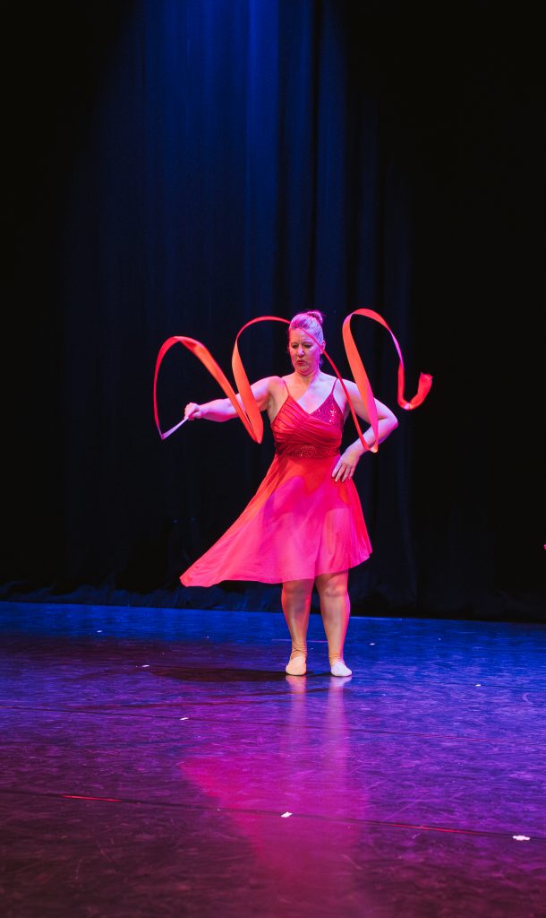 Female dancer in a bright pink dress performing a ribbon dance on a dark stage with blue lighting, ribbons swirling around.