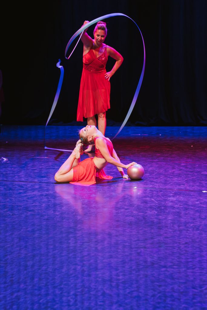 Two female performers in red costumes on a dark stage, one standing with a hoop raised above her, the other lying on the floor with a hoop nearby and a ball nearby.