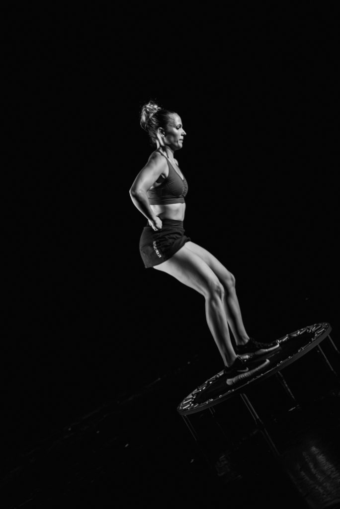 Black-and-white photo of a woman in athletic wear mid-jump on a trampoline against a dark background.