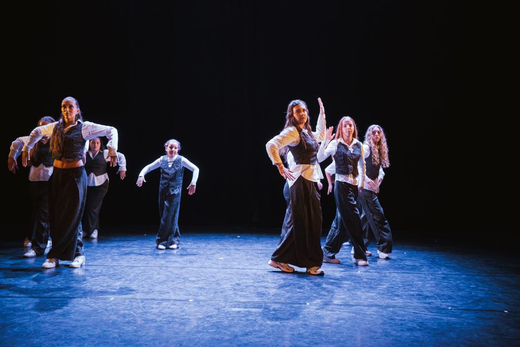 Group of dancers in white shirts and pinstripe vests performing a routine on a dark stage with blue lighting.
