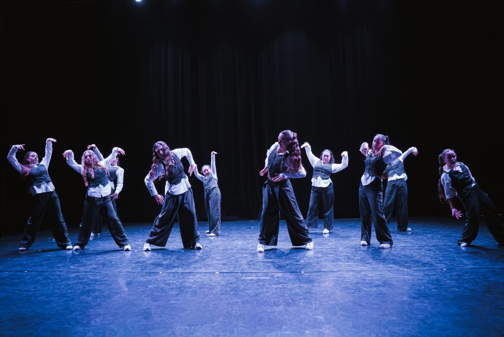 Group of dancers in matching striped outfits perform on a dark stage with blue lighting.