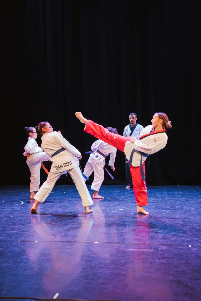 Martial arts students practicing kicks on a stage with dark curtains; one student in red uniform delivers a high kick while others watch.