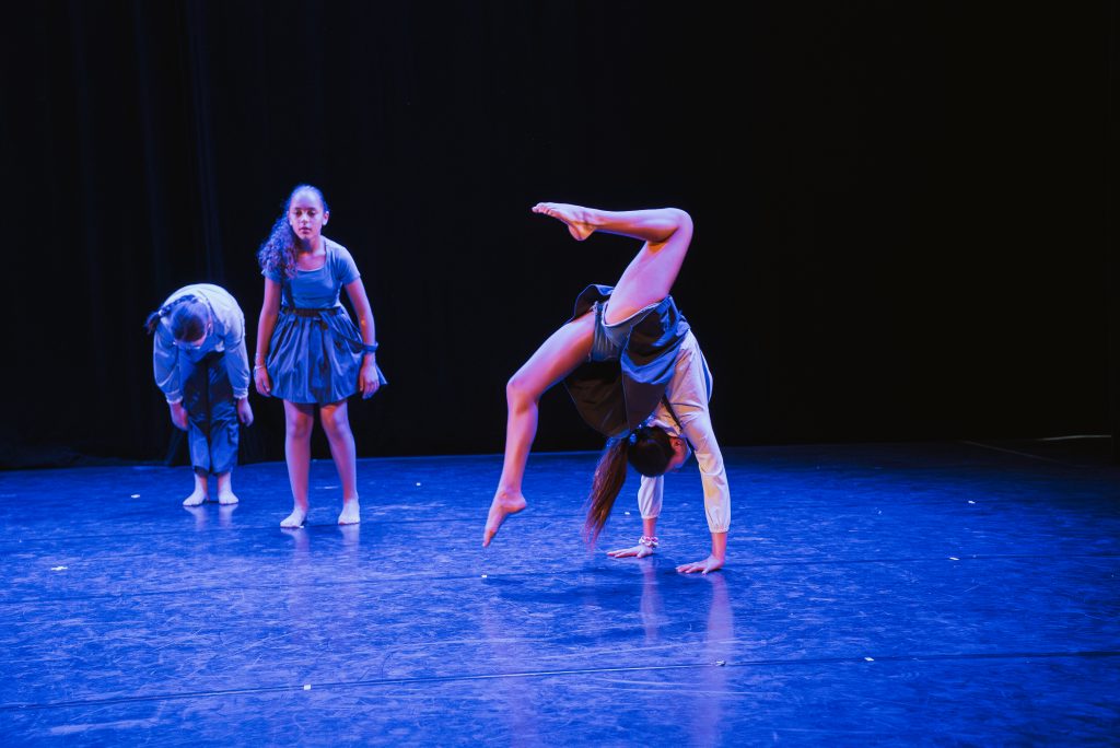 Group of young dancers in blue costumes on a dark stage; one dancer is performing a handstand while others look on.