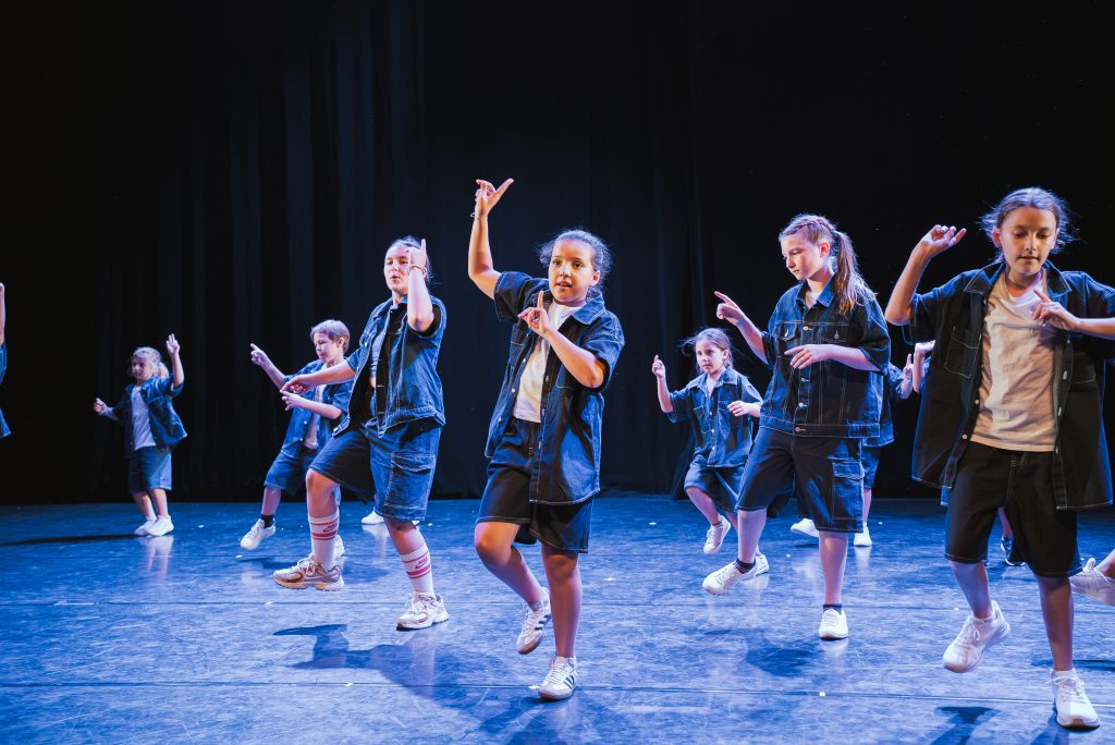 Group of children dancing on a stage, wearing denim jackets and shorts, mid-movement under blue stage lights