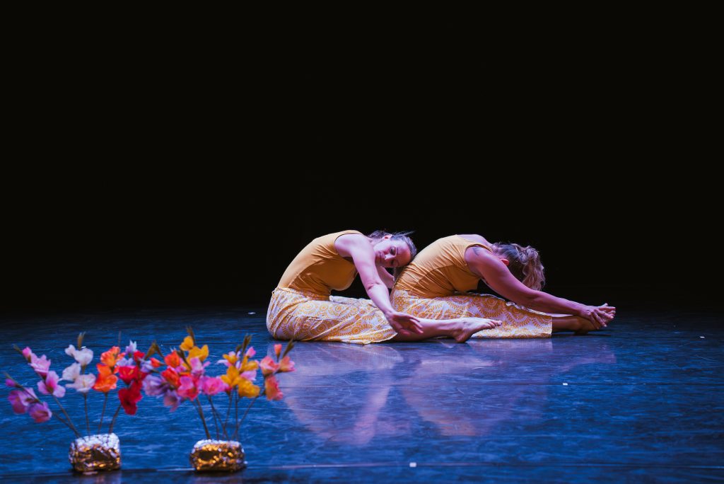 Two dancers in orange outfits bend forward in a synchronized floor pose on a dim stage, with colorful flowers in the foreground.