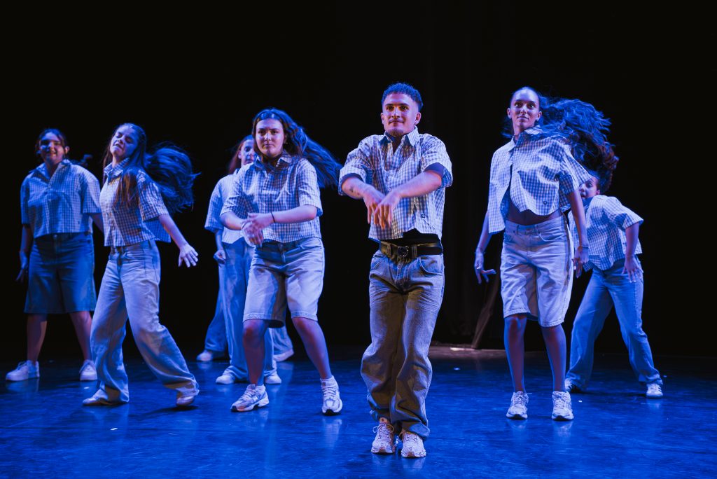Group of young dancers performing on stage in matching plaid shirts and light jeans under blue stage lighting