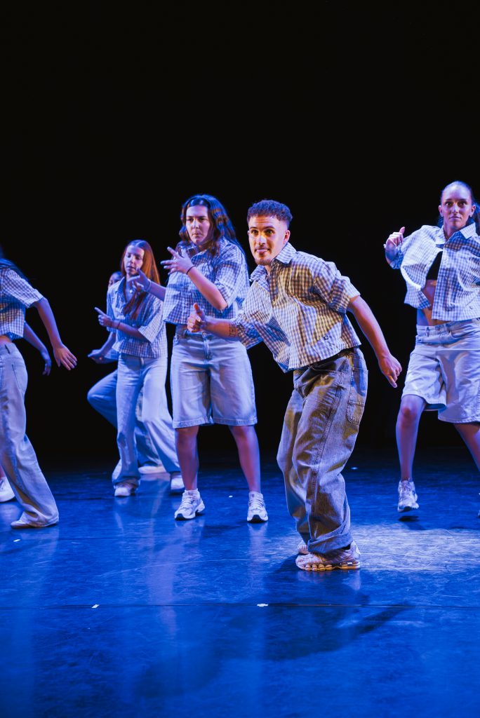 Group of young dancers in plaid shirts and jeans performing a choreographed routine on a dark stage with blue lighting