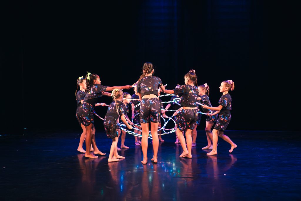 Group of young girls in colorful, splatter-pattern dance outfits twirling ribbons on a dark stage.