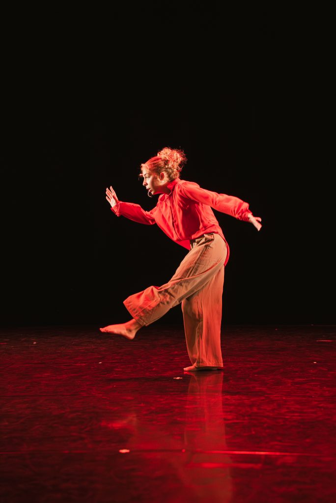 Female dancer in red outfit performs a dynamic pose on stage, one leg extended and arms outstretched against a dark background.