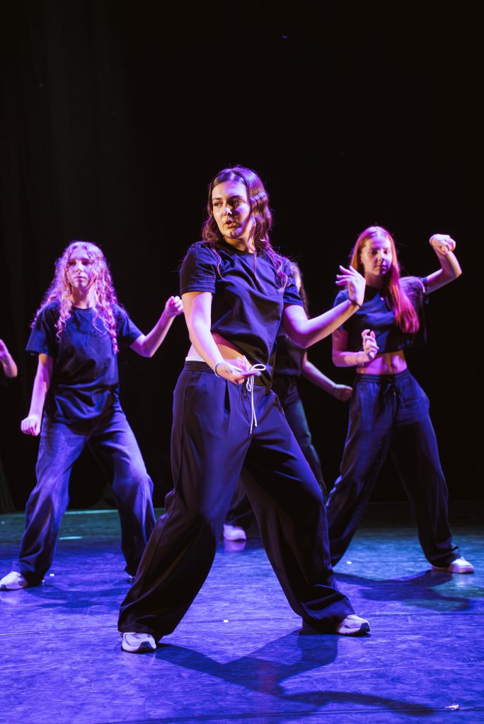 Dancers performing on a stage in black outfits under blue stage lighting, with a central female dancer in the foreground.