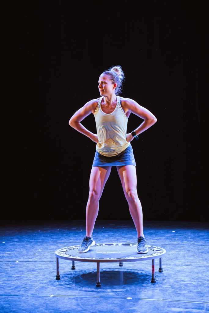 Woman in athletic wear stands with hands on hips on a small trampoline on a dark stage.