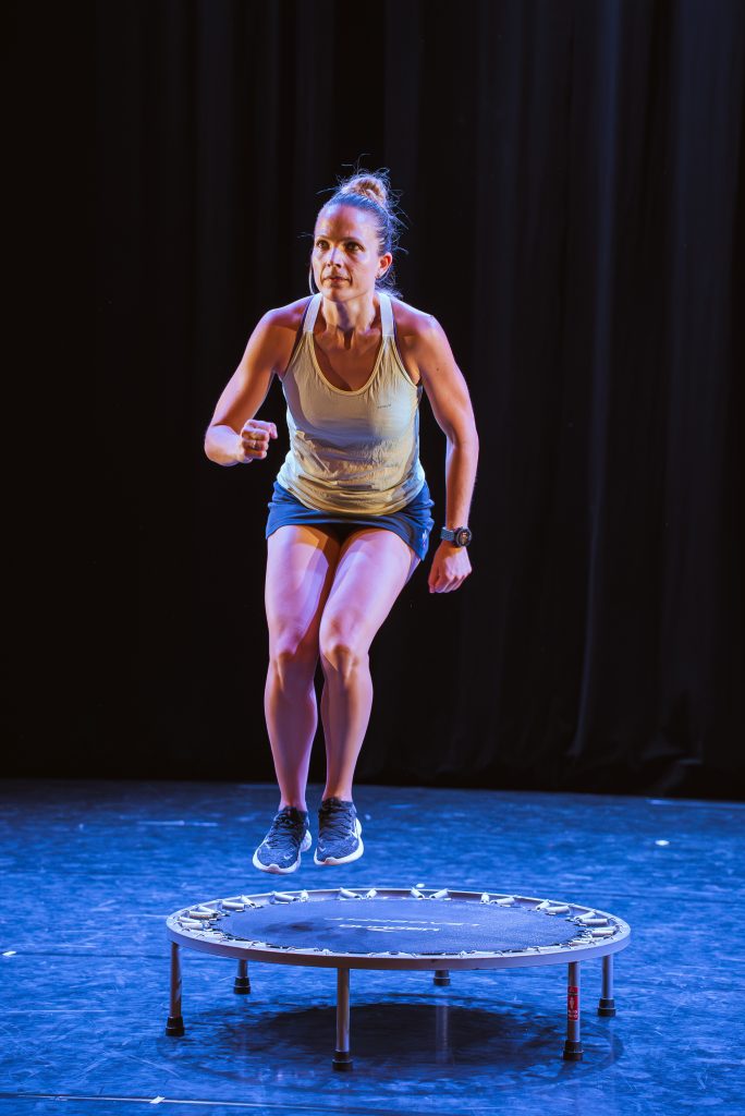 Female athlete in a white tank top and blue shorts mid-jump on a trampoline on a dimly lit stage with a dark curtain backdrop.