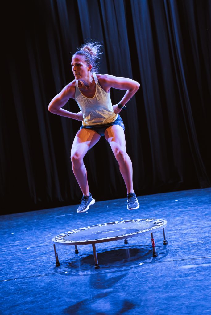 Female athlete mid-jump on a small trampoline on a stage, wearing a white tank top and dark shorts with blue lighting behind.