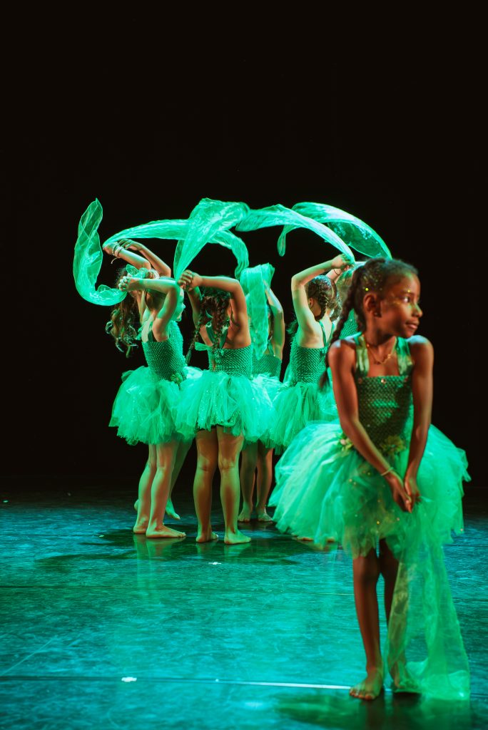 Group of young dancers in turquoise costumes with flowing green fabric on stage, facing in a circle against a dark background.