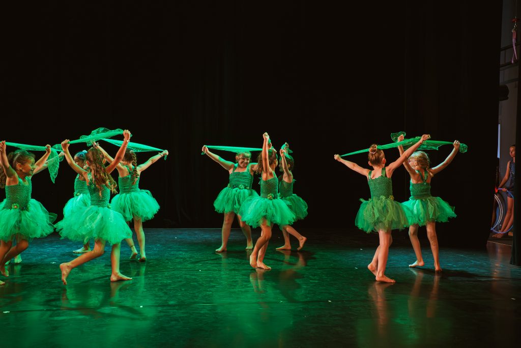 Group of young dancers in green tutus holding green scarves on stage in a coordinated circle formation.