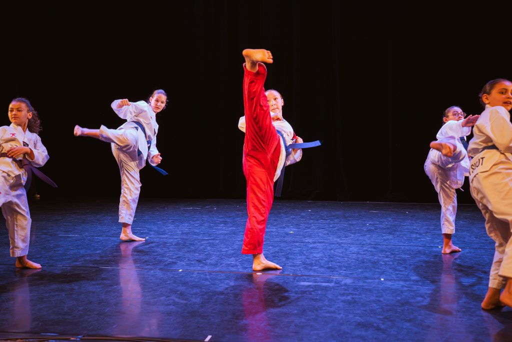 Group of children in white martial arts gis with blue belts performing a high kick on a dark stage; central performer wears a red uniform.