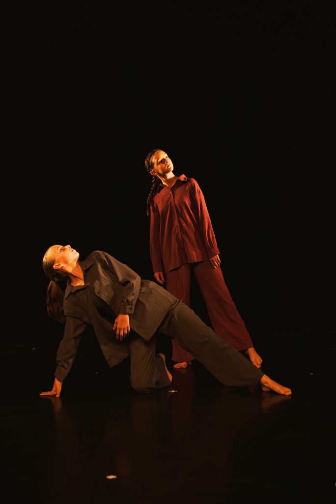 Two dancers perform on a dark stage: one standing in a burgundy outfit looking upward, the other crouched and leaning on the floor in a gray outfit under warm lighting.