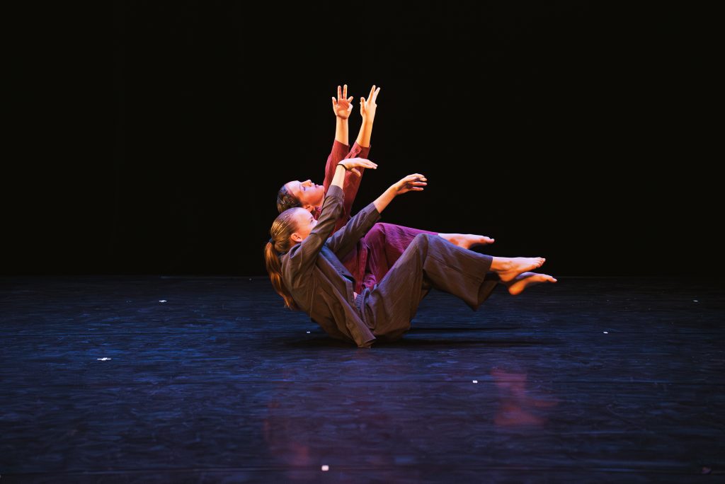 Three dancers lift their bodies and arms in a synchronized contemporary dance against a dark stage with minimal lighting on the performers.