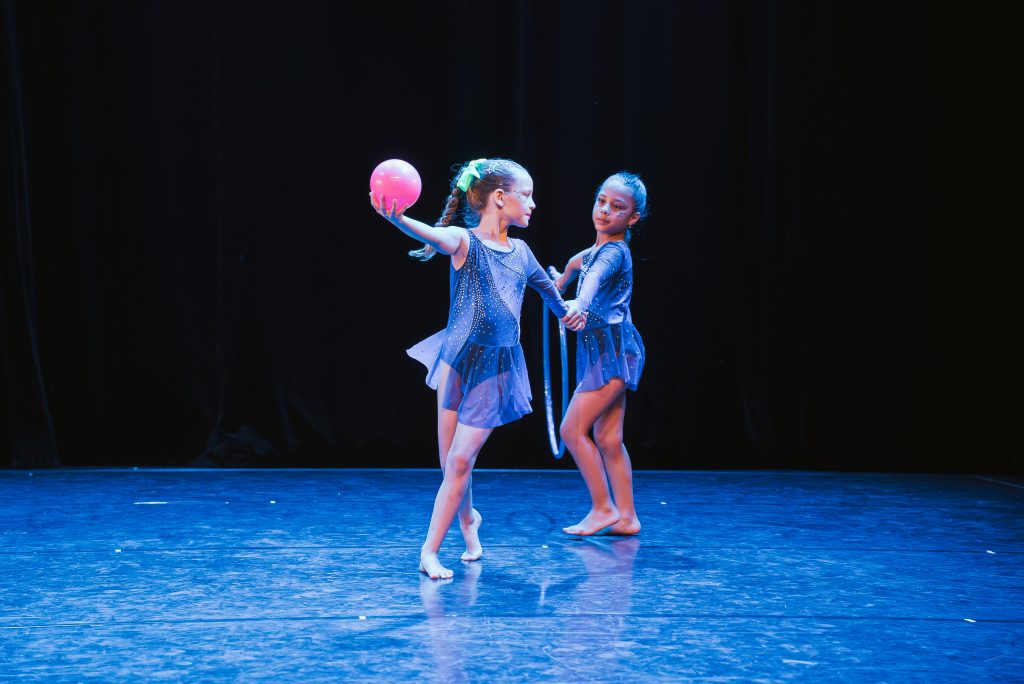 Two young girls in blue sparkly dance costumes perform on a dark stage, one holding a pink ball as they pose mid-performance.