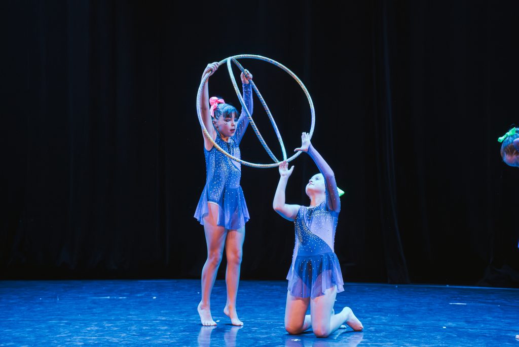 Young gymnasts in blue costumes perform with sparkling hoops on a dark stage.