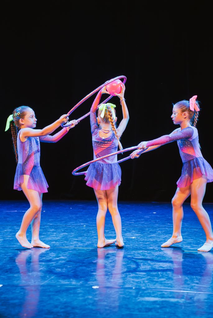 Four young girls in purple sparkly costumes perform a hoop-dance on a dark stage; one holds a pink ball above her head.