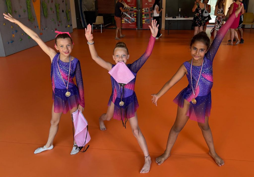Three young gymnasts in purple-pink leotards raise their arms in celebration, each wearing a medal on an orange gym floor.