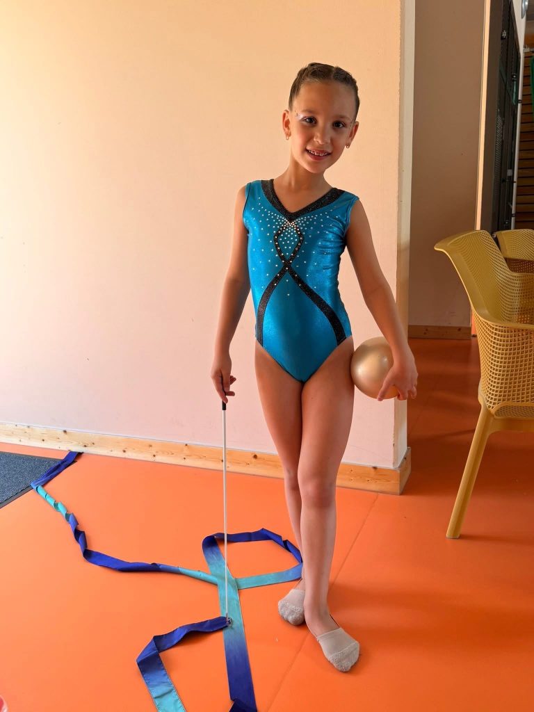Young girl in a blue leotard stands on an orange gym mat, holding a baton with a blue ribbon trailing on the floor.
