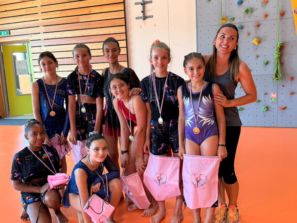 Group of young gymnasts wearing leotards with medals posing with their coach in a gym with a climbing wall background.