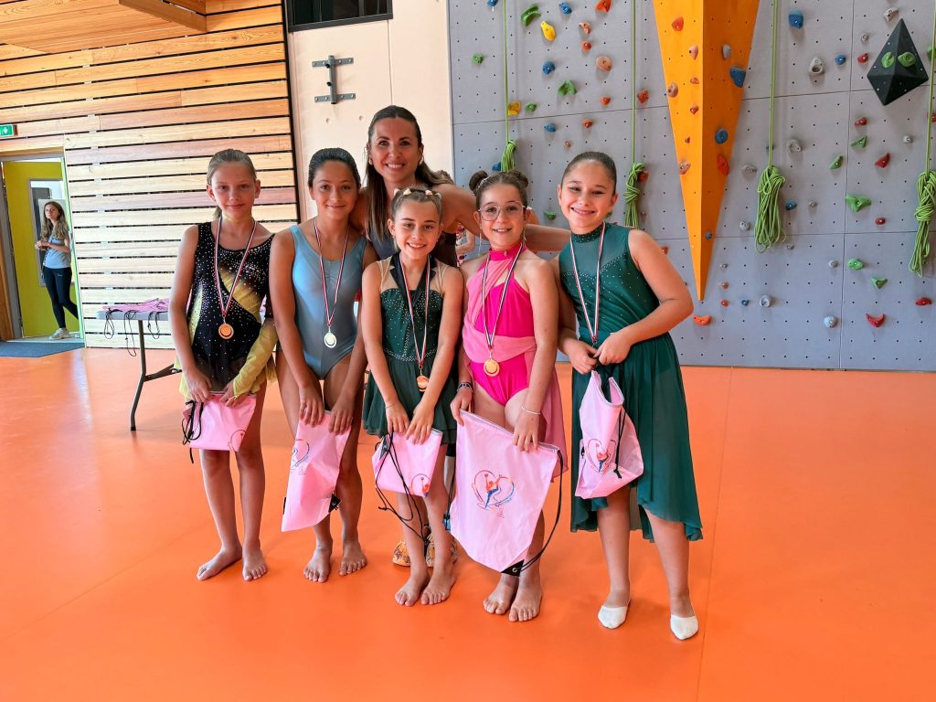 Five young gymnasts in colorful leotards with medals pose for a group photo with their coach in a gym featuring a climbing wall in the background.