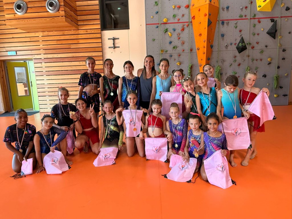 Group of young girls in colorful athletic outfits posing with medals and pink bags indoors, gymnasium backdrop with climbing wall.