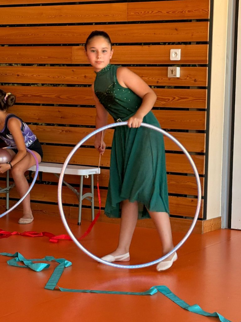 Young performer in a green dress stands with a silver hula hoop in a gym, wooden wall panels in the background.