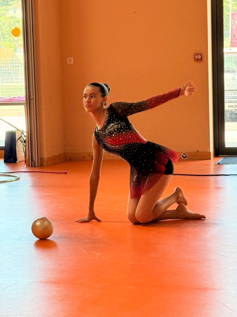 Gymnast on an orange floor performing a pose with the right arm extended backward, left hand on the floor, with an orange ball nearby.