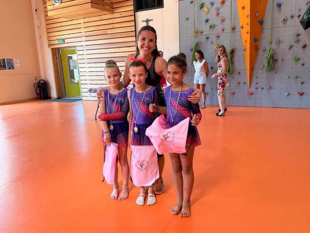 Three young gymnasts in matching purple leotards with medals pose with their coach in a gym setting, pink bags in hand.