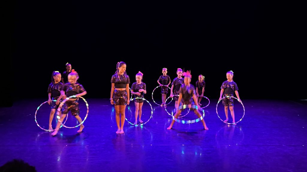 Group of young dancers in dark costumes with colorful hoops performing on a stage.