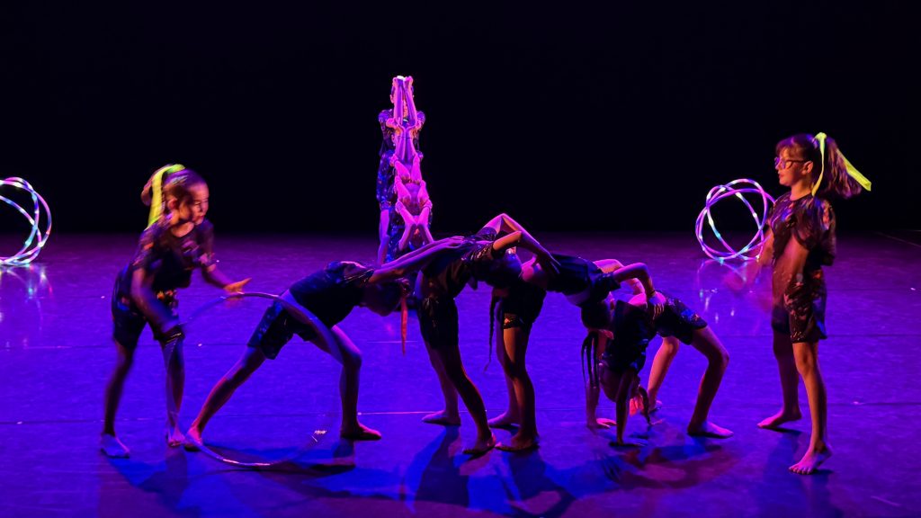 Dance troupe performing on a dim purple-lit stage with glowing LED hoops and coordinated movements.