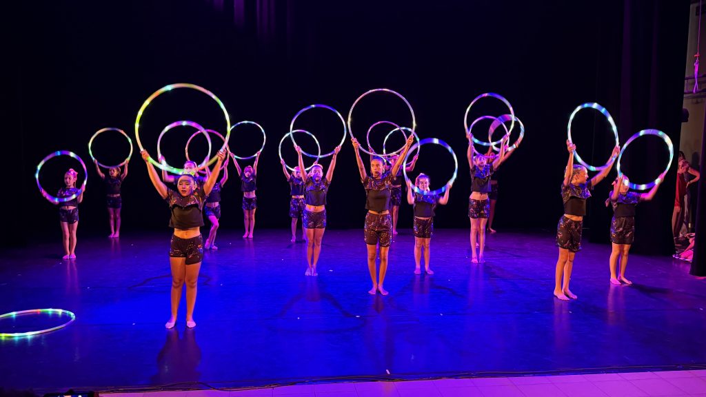 A group of young performers on a dark stage spinning illuminated LED hoops in rainbow colors, forming circles overhead and around them.
