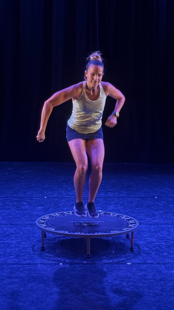Woman mid-jump on a small round trampoline on a stage, wearing a light tank top and dark shorts under blue stage lighting.