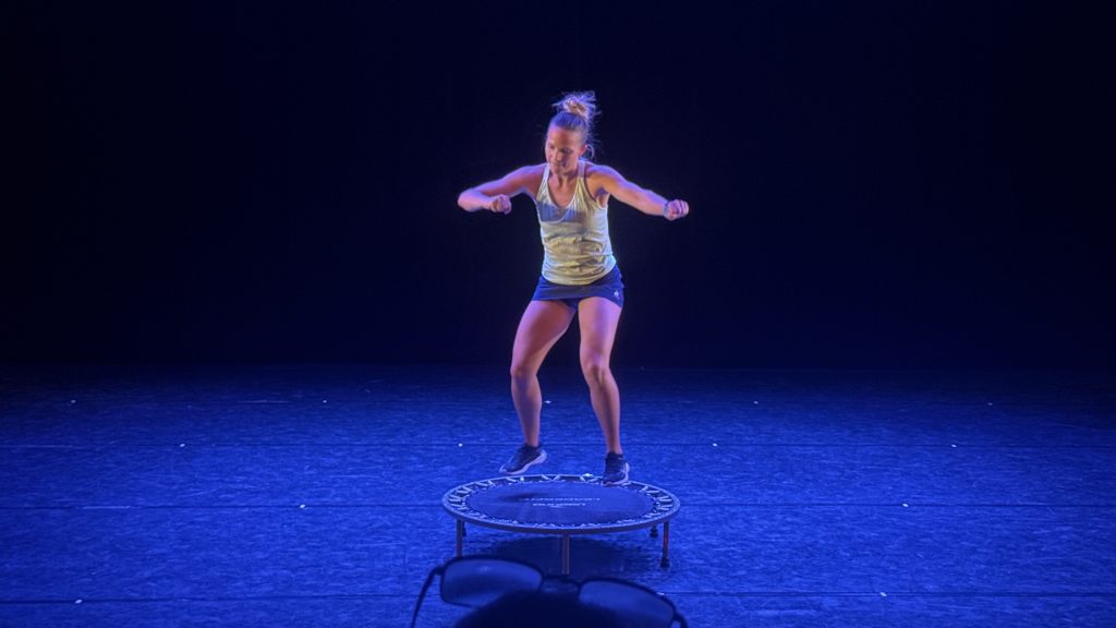 Woman in a light tank top and shorts bouncing on a mini-trampoline on a dark stage with blue lighting.
