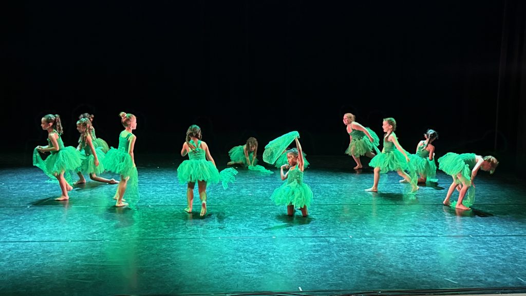Young girls in bright green tutus performing a ballet on a dark stage.