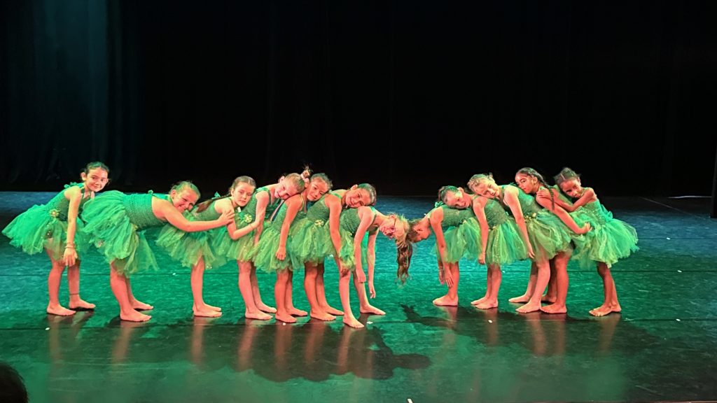 Group of young dancers in bright green tutus bowing together on stage after a performance.