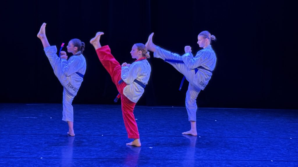 Four martial artists perform high kicks in a row on a stage; three wear white gis with blue belts, one wears a red gi, against a dark backdrop.